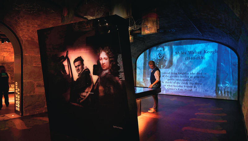 Man in museum interacts with display about Myles Walter Keogh, historical text projected on arched wall.
