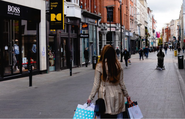 Person walking down a shopping street with bags, wearing a beige coat.