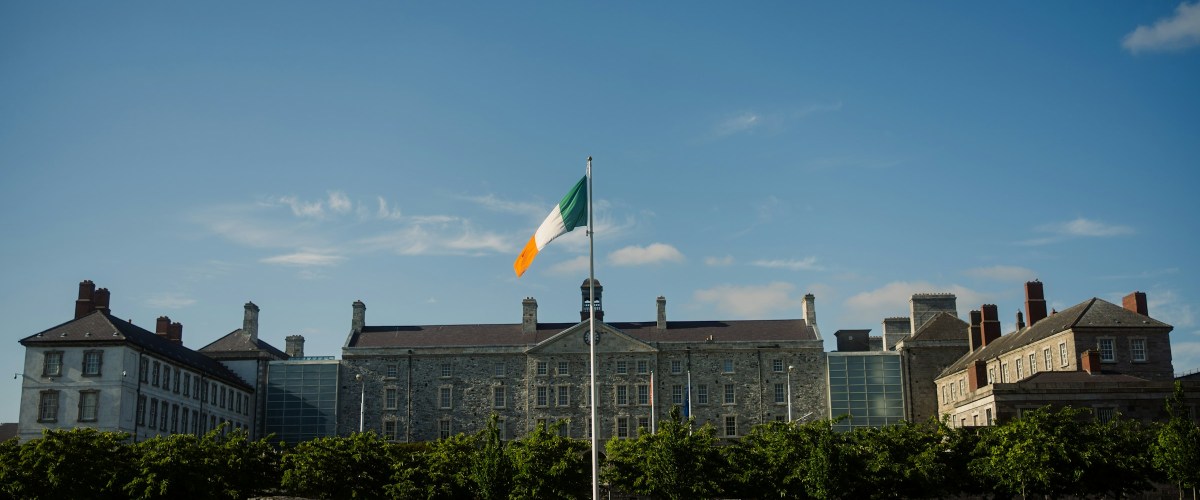 roger-nobles-wcsx7p6Owjk-unsplash Stone building and Irish flag under blue sky with trees and grassy foreground.