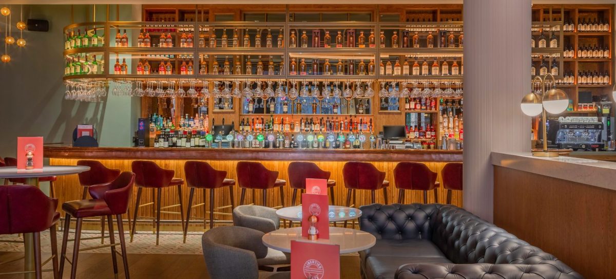 Modern bar interior with red velvet chairs, marble tables, and fully stocked shelves behind the counter.